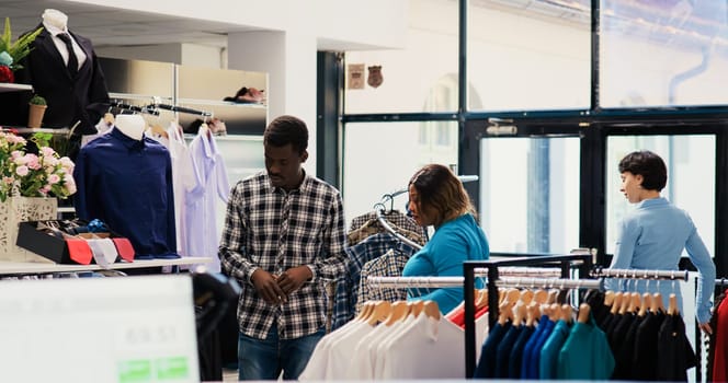 African american couple shopping for formal wear, checking trendy clothes fabric before buying it in modern boutique. Stylish people looking at hangers with new fashion collection in clothing store