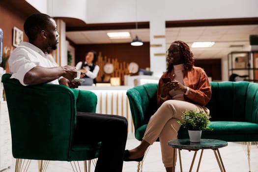 Couple sitting in lounge area at resort, waiting to fill in registration at reception front desk. Young man and woman preparing for check in, relaxing on couch in hotel lobby.