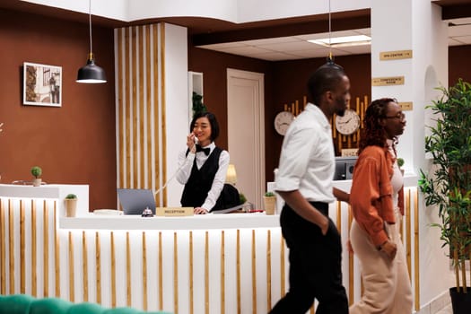 Asian receptionist making online booking using landline phone at reception counter front desk, talking to people about room reservations. Hotel staff working on welcoming tourists.