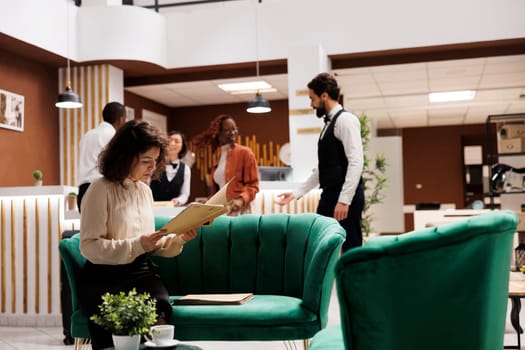 Hotel manager checking record files in lobby lounge area, reviewing registration forms and bureaucracy reports. Young resort administrator sitting on couch to ensure excellent service.