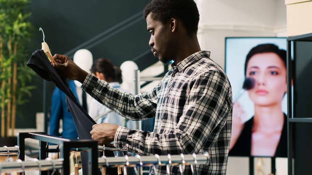 Stylish customer shopping for casual wear, analyzing merchandise fabric in modern boutique. African american man looking at hangers with new fashion collection, checking basic outfits
