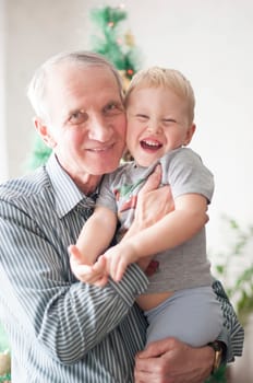 handsome gray-haired Caucasian grandfather hugs a little boy grandson against the background of a New Year tree in a warm house in a close family circle, home sweet home, High quality photo