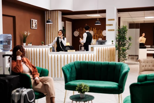 Asian employee answering booking call, using landline phone to write new room registration at hotel reception desk. Hotel worker providing excellent service to future guests, working at resort.