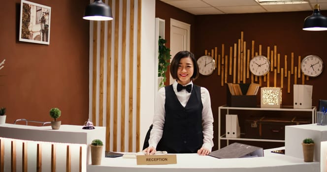 Happy professional asian hotel employee taking phone calls while working at reception counter. Joyful smiling receptionist taking travel accommodation booking requests standing at check in front desk in luxurious lobby