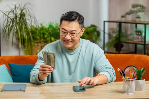 Planning budget. Rich happy Asian man use smartphone calculator app counting money cash at table desk home room apartment. Guy satisfied of income earnings win saves money for planned vacation gifts