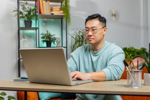 Confident concentrated Asian man working remotely on a project on laptop computer, thinking, typing on keyboard at home apartment. Freelancer guy communicates online chat sits on couch at table desk