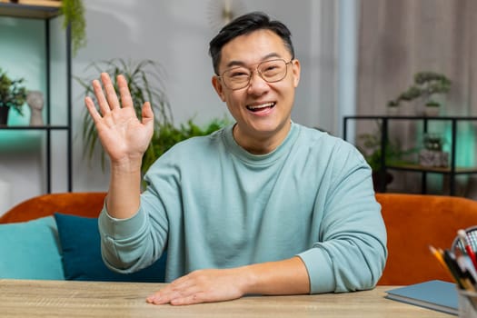 Hello. Asian handsome man smiling friendly at camera and waving hands gesturing hello hi, greeting or goodbye welcoming with hospitable expression at home. Chinese guy sitting on sofa in living room