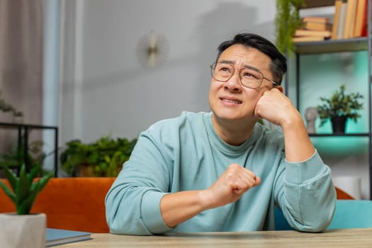 Portrait of sad Asian man guy sitting at home table looks pensive thinks over life concerns or unrequited love, suffers from unfair situation. Problem, break up, depressed feeling bad annoyed, burnout
