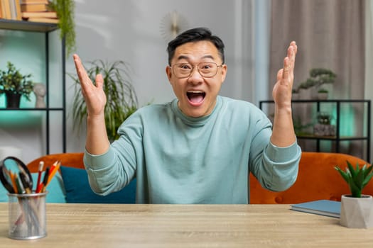Happy Asian man shouting, celebrating success, winning, goal achievement good news lottery luck victory, watching TV championship at modern home apartment. Chinese guy in living room sits on couch
