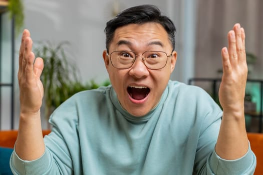 Surprised happy Asian man shouting, celebrating success, winning, goal achievement good news lottery luck victory, watching TV championship at modern home apartment. Chinese guy in room sits on couch