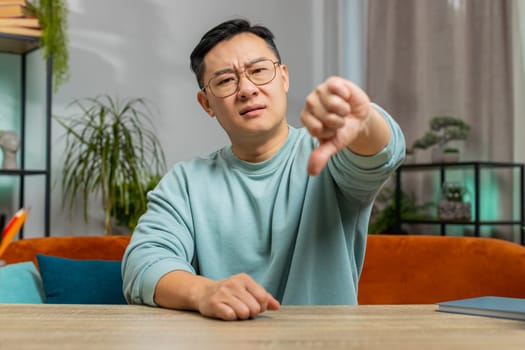 Dislike. Upset unhappy middle-aged Chinese man showing thumbs down sign gesture, expressing discontent disapproval dissatisfied bad work at home apartment indoors. Displeased Asian guy in room on sofa