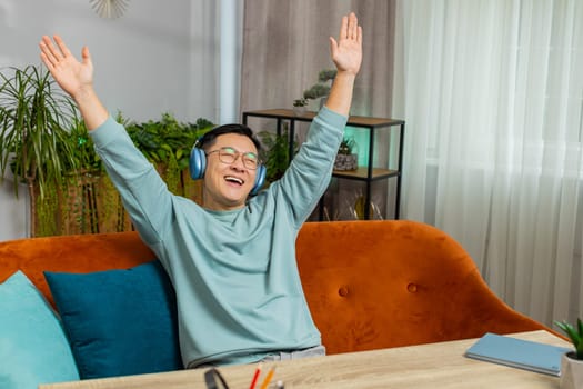 Happy relaxed overjoyed mature Asian man in wireless headphones choosing listening favorite energetic disco music, dancing raises hands entertaining on couch at home. Chinese people weekend activities