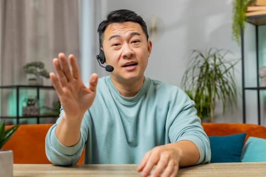 Asian Chinese man wearing headset, freelance worker call center or support service operator helpline. Chinese guy having talk with client or colleague communication support waving hello alone at home