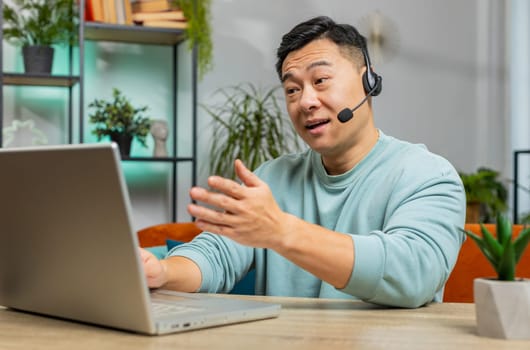 Asian man wearing headset, freelance worker, call center or support service operator helpline, having talk with client or colleague communication support at home room. Guy sits on couch at table desk