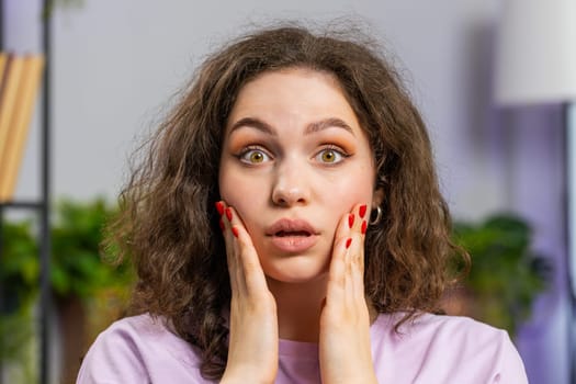 Oh my God, Wow. Young woman surprised looking at camera with big eyes, shocked by sudden victory, good win news, celebrating at home room. Excited amazed adult girl indoors in living room
