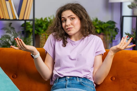 I dont know what to say. Confused pretty woman feeling embarrassed about ambiguous question, having doubts, no idea, being clueless and uncertain at home. Portrait of girl sits on sofa in living room