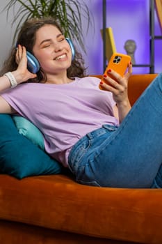 Happy overjoyed woman in wireless headphones relaxing lying on couch at home apartment choosing listening favorite energetic disco dancing music. People weekend daytime leisure activities. Vertical