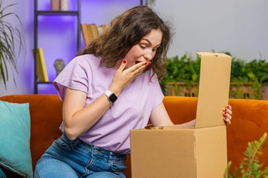 Happy young woman unpacking delivery parcel sitting at home apartment on sofa. Smiling satisfied girl shopper online shop customer opening cardboard box receiving purchase gift by fast postal shipping