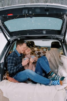 Smiling couple sitting on blanket in car trunk with mugs of coffee in winter forest. High quality photo