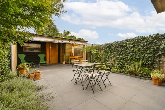 a patio with tables and chairs in the middle of it, surrounded by lush green plants on either side of the house