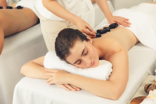Hot stone massage at spa salon in luxury resort with day light serenity ambient, blissful couple customer enjoying spa basalt stone massage glide over body with soothing warmth. Quiescent