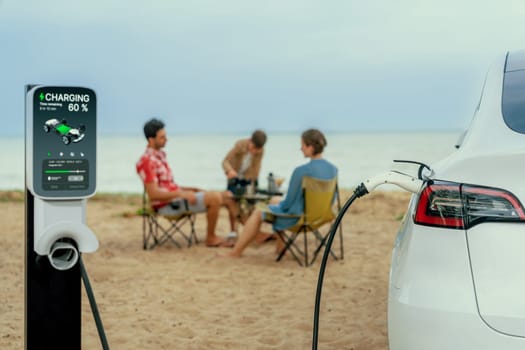 Alternative family vacation trip traveling by the beach with electric car recharging battery from EV charging station with blurred family enjoying the seascape background. Perpetual