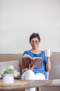 A happy retired 60s Asian woman reading a book while having her coffee on a sofa at home..