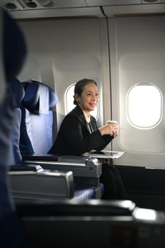 Charming middle aged businesswoman drinking coffee and looking away on the airplane during flight.