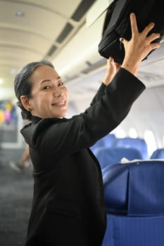 Smiling middle age woman passengers putting her luggage into the overhead compartment on an airplane.