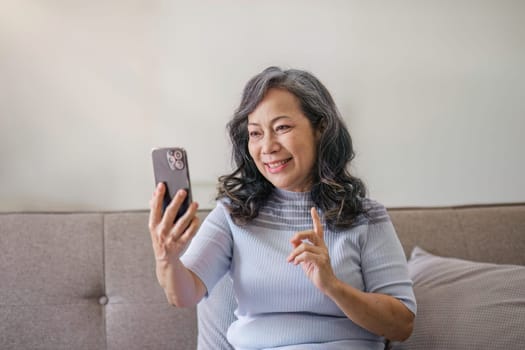 Portrait, Happy 60s retired Asian woman using her phone on a sofa in the living room. chatting, scrolling on the phone.