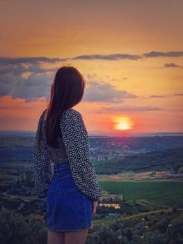 Rear view of a girl watching the sunset from the top over the valley. Natural summer evening scene