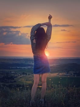 Wild and free youth concept with a relaxed girl, rear view stands with hands up in the wind facing the beautiful summer sunset on the top of a hill looking over the valley