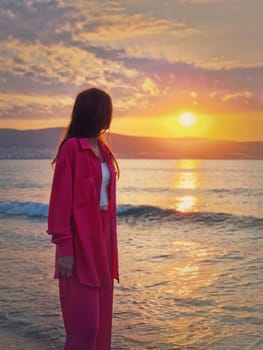 Aesthetic young woman portrait wearing pink suit on the sea shore as meets the dawn. Beautiful sunrise at the beach. Summer vacation background, holiday relaxation concept