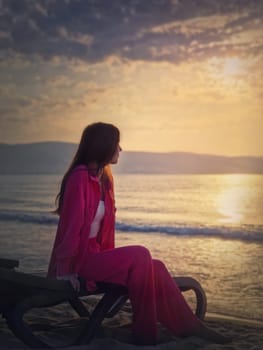 Carefree young woman aesthetic portrait sitting relaxed on the sunbed at the beach looking at the sunrise above the sea. Beautiful seaside dawn scene. Summer holiday, travel and relaxation concept