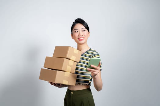 Young happy asian woman wear summer casual clothes hold stack cardboard blank boxes use mobile cell phone 