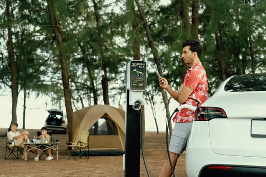 Outdoor adventure and family vacation camping in nature travel by eco friendly car for sustainable future. Lovely sit on trunk, charging EV car with EV charging station in campsite. Perpetual