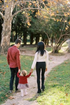 Parents with a little girl walk along the path in the park holding hands. Back view. High quality photo