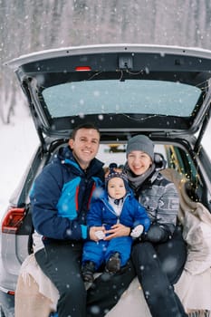 Smiling mom, dad and little girl sitting on blanket in car trunk in winter forest. High quality photo