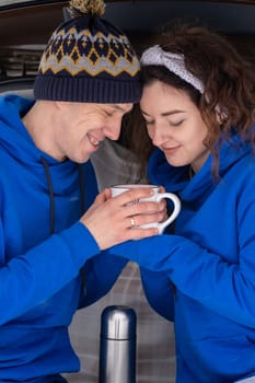 Outdoor happy couple in love posing in cold winter weather. A man and a woman in blue hoodies. Emotional young couple having fun while walking by winter forest, loving man hugging his laughing woman