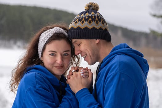 Outdoor happy couple in love posing in cold winter weather. A man and a woman in blue hoodies. Emotional young couple having fun while walking by winter forest, loving man hugging his laughing woman