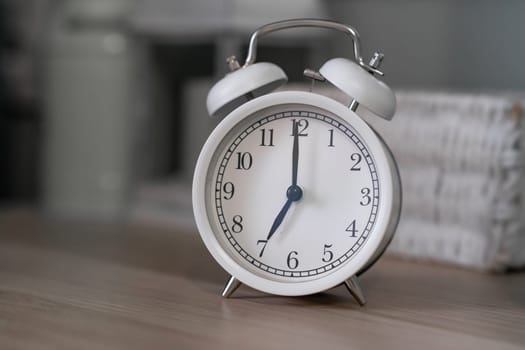 Close-up of a round white alarm clock on a table in the bedroom. The hands on the clock show seven o'clock in the morning, time to get up. Retro alarm clock on the table, vintage tone. space for text