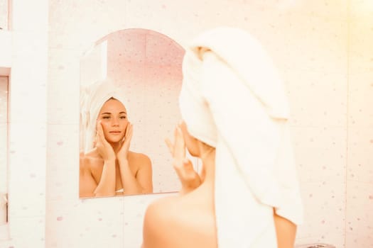 Young beautiful woman using face cream moisturizing lotion after bath. Pretty attractive girl in a towel on her head stands in front of a mirror in a home bathroom. Daily hygiene and skin care.