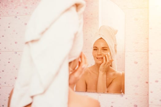 Young beautiful woman using face cream moisturizing lotion after bath. Pretty attractive girl in a towel on her head stands in front of a mirror in a home bathroom. Daily hygiene and skin care.