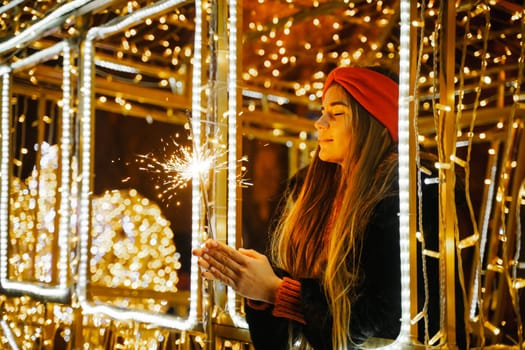 Woman holding sparkler night while celebrating Christmas outside. Dressed in a fur coat and a red headband. Blurred christmas decorations in the background. Selective focus.