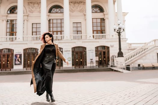 Woman street lifestyle. Image of stylish woman walking through European city on sunny day. Pretty woman with dark flowing hair, dressed in a beige raincoat and black, walks along the building