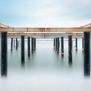 Fine art view of columns under metal pier at sunrise. Shot with long exposure to make the sea appear as fog