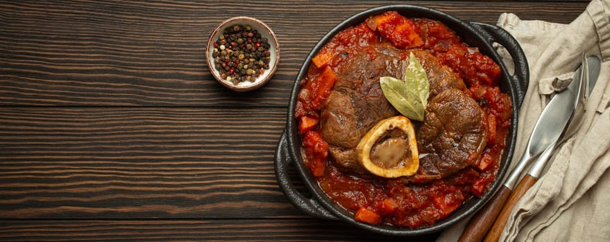 Traditional Italian dish Ossobuco all Milanese made with cut veal shank meat with vegetable tomato sauce served in black casserole pan top view on rustic brown wooden background, copy space.