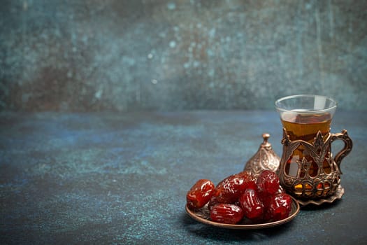 Breaking fasting with dried dates during Ramadan Kareem, Iftar meal with dates and Arab tea in traditional glass, angle view on rustic blue background. Muslim feast, space for text.