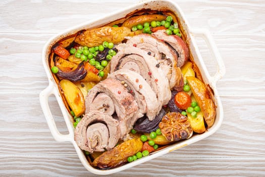 Rolled sliced pork roasted in white casserole dish with potatoes, vegetables and herbs on rustic white wooden background top view. Baked pork roll with vegetables for dinner.