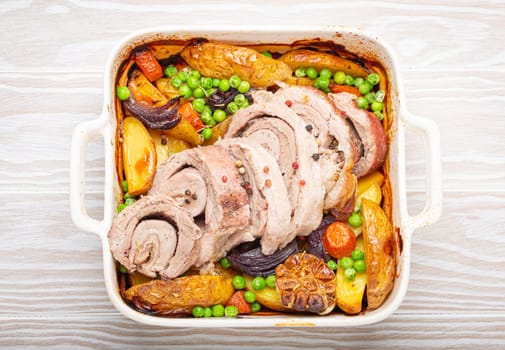 Rolled sliced pork roasted in white casserole dish with potatoes, vegetables and herbs on rustic white wooden background top view. Baked pork roll with vegetables for dinner.